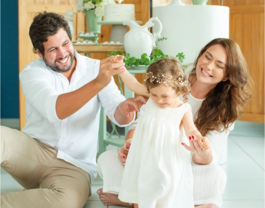 Pais sentados no chão, ajudando sua filhinha a andar, em sua festa de batizado, todos muito sorridentes.