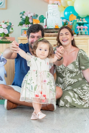 Clara e os pais na frente da mesa do bolo, com seus pais sentado e o pai lhe segurando pelos braços a ajudando a andar.