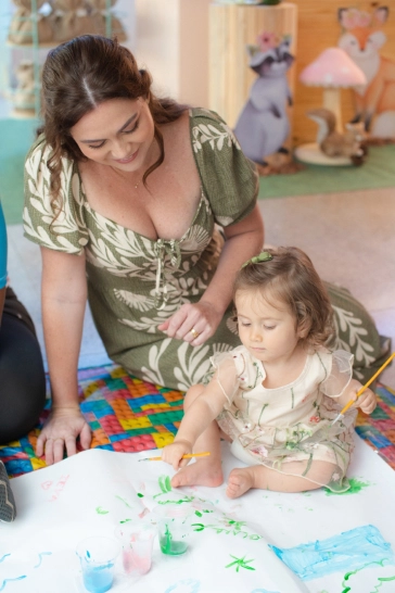 Clara e sua mãe no chão, Clara segura pincéis e uma arte com tintas numa grande folha branca.