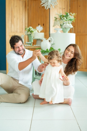 Pais sentados no chão, ajudando sua filhinha Clara a andar, em sua festa de batizado, todos muito sorridentes.