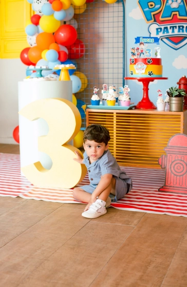 Lucas sentado na frente da mesa do bolo, apoiado no grande número três que faz parte da decoração.