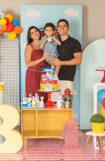 Lucas e seus pais, todos sorrindo, posando para uma foto na frente da mesa do bolo.